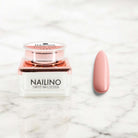NAILINO Farbgel Coral Kiss in Glastiegel mit Nagel in sanften Korallton auf marmoriertem Hintergrund